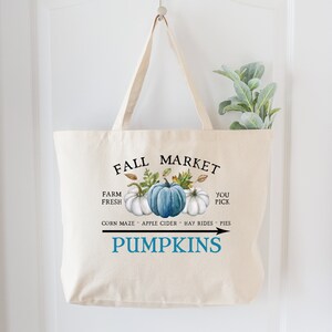 Op de afbeelding: Een witte canvas draagtas met een blauw en wit pompoenontwerp. De tekst "Fall Market" is in het zwart bovenaan de tas gedrukt. De tekst "Farm Fresh" is in het zwart aan de linkerkant van de tas gedrukt. De tekst "You Pick" is in het zwart aan de rechterkant van de tas gedrukt. De tekst "Corn Maze - Apple Cider - Hay Rides - Pies" is in het zwart onder de pompoenen gedrukt. De tekst "Pumpkins" is in het blauw onder de tekst gedrukt. Een pijl wijst naar rechts.