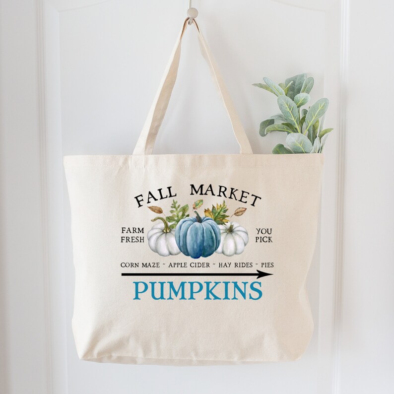 Op de afbeelding: Een witte canvas draagtas met een blauw en wit pompoenontwerp. De tekst "Fall Market" is in het zwart bovenaan de tas gedrukt. De tekst "Farm Fresh" is in het zwart aan de linkerkant van de tas gedrukt. De tekst "You Pick" is in het zwart aan de rechterkant van de tas gedrukt. De tekst "Corn Maze - Apple Cider - Hay Rides - Pies" is in het zwart onder de pompoenen gedrukt. De tekst "Pumpkins" is in het blauw onder de tekst gedrukt. Een pijl wijst naar rechts.