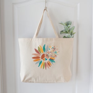 May include: A beige canvas tote bag with a colorful peace sign sunflower design. The sunflower is made up of peace signs and has a variety of colors including orange, yellow, green, blue, and red.