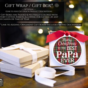 May include: A white ceramic ornament with a red and green plaid pattern and the text "Merry Christmas to the Best Papa Ever". The ornament is in a white gift box with a red ribbon.