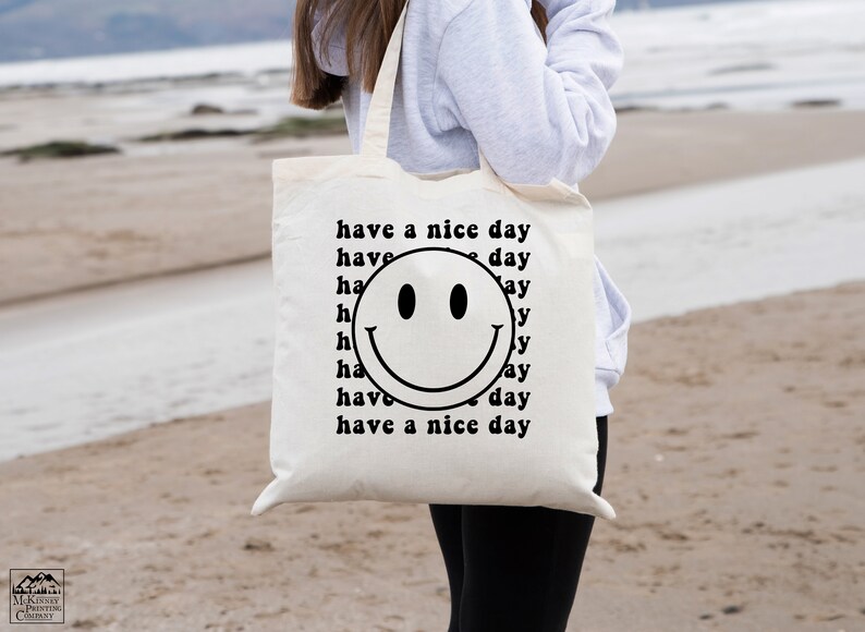 May include: White canvas tote bag with a black smiley face and the text "have a nice day" repeated around the smiley face.