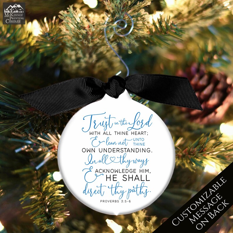 May include: White Christmas ornament with a black ribbon and silver hanger, inscribed with a religious quote in blue script. The ornament is hanging on a Christmas tree. The text reads "Trust in the Lord..." and "Customizable message on back."