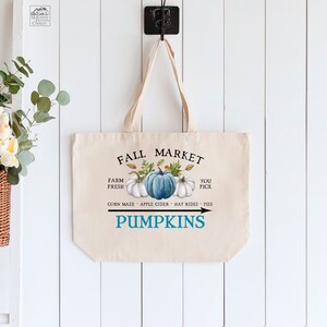 Op de afbeelding: Een witte canvas draagtas met een blauw en wit pompoenontwerp en de tekst "Fall Market" en "Pumpkins". De tekst zegt ook "Farm Fresh" en "You Pick" met een lijst van activiteiten: "Corn Maze, Apple Cider, Hay Rides, Pies".