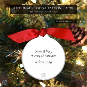 May include: White Christmas ornament with a red ribbon bow, hanging on a Christmas tree. The ornament has the text "Have A Very Merry Christmas!! -Olivia 2029". The background is a blurred image of a Christmas tree with lights.