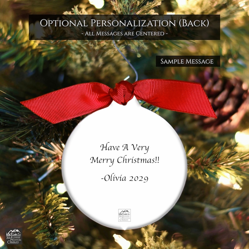 May include: White Christmas ornament with a red ribbon bow, hanging on a Christmas tree. The ornament has the text "Have A Very Merry Christmas!! -Olivia 2029". The background is a blurred image of a Christmas tree with lights.