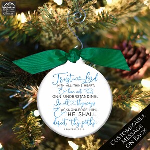 May include: White Christmas ornament with a green ribbon and silver hanger. The ornament features the text "Trust in the Lord" and a Bible verse. The background shows a Christmas tree with pine needles and a pine cone.