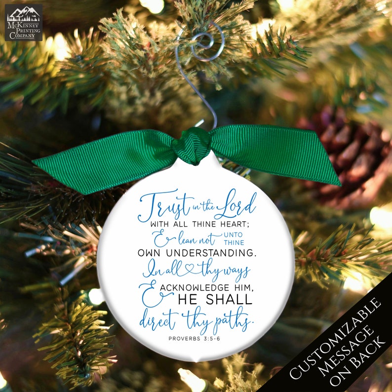 May include: White Christmas ornament with a green ribbon and silver hanger. The ornament features the text "Trust in the Lord" and a Bible verse. The background shows a Christmas tree with pine needles and a pine cone.