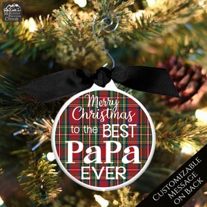 May include: A round white Christmas ornament with a red and green plaid pattern. The ornament has the text "Merry Christmas to the Best Papa Ever" in white lettering. The ornament is hanging on a Christmas tree with a black ribbon.