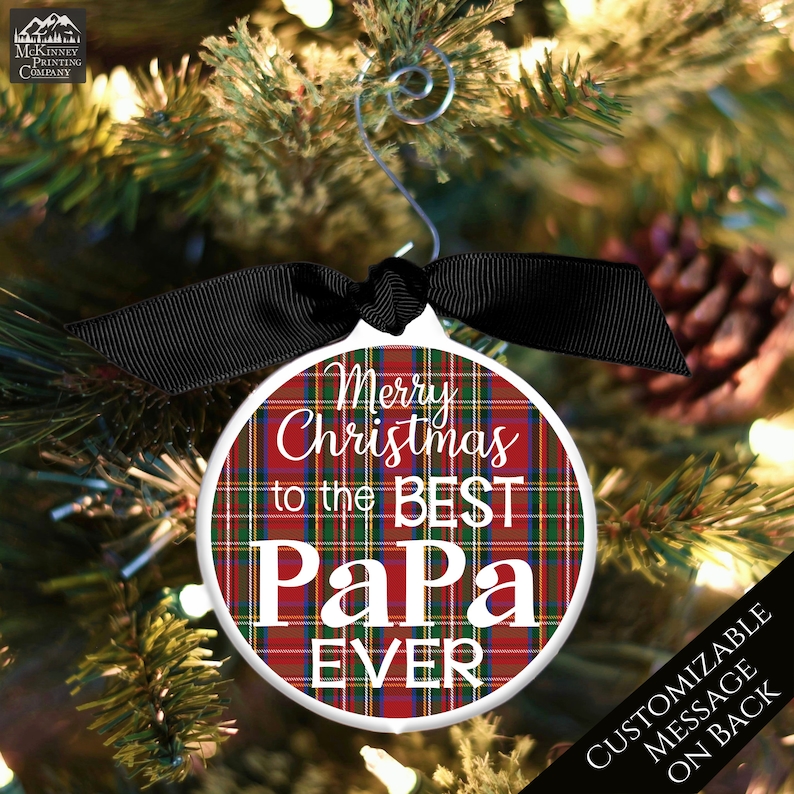 May include: A round white Christmas ornament with a red and green plaid pattern. The ornament has the text "Merry Christmas to the Best Papa Ever" in white lettering. The ornament is hanging on a Christmas tree with a black ribbon.