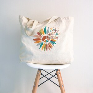 May include: A white canvas tote bag with a colorful peace sign design. The design features a peace sign surrounded by stylized flower petals in shades of orange, yellow, green, and blue. The peace sign is repeated in smaller sizes around the flower.