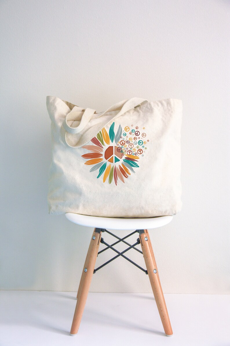 May include: A white canvas tote bag with a colorful peace sign design. The design features a peace sign surrounded by stylized flower petals in shades of orange, yellow, green, and blue. The peace sign is repeated in smaller sizes around the flower.