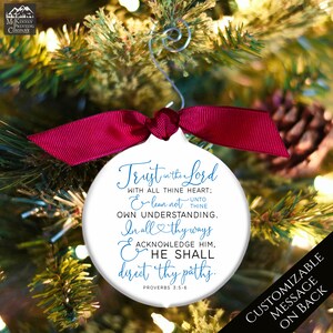 May include: White Christmas ornament with a red ribbon and silver hanger. The ornament has the text "Trust in the Lord" and a Bible verse. The background is a Christmas tree.