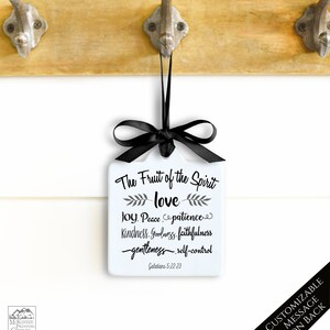 May include: A white square ornament with a black ribbon and bow, featuring the text "The Fruit of the Spirit" and related words. The ornament hangs from a wooden surface with metal hooks. The text "CUSTOMIZABLE MESSAGE ON BACK" is also visible.