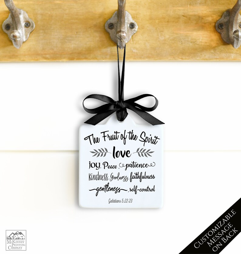 May include: A white square ornament with a black ribbon and bow, featuring the text "The Fruit of the Spirit" and related words. The ornament hangs from a wooden surface with metal hooks. The text "CUSTOMIZABLE MESSAGE ON BACK" is also visible.