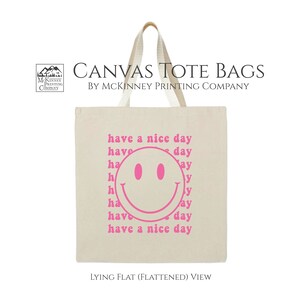 May include: A white canvas tote bag with pink text that says "have a nice day" and a pink smiley face. The bag is lying flat.