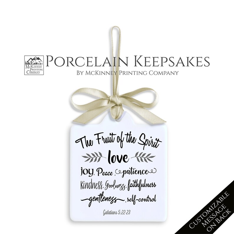May include: A white porcelain keepsake with a beige ribbon and bow. The text "The Fruit of the Spirit" is above the word "love", followed by other words. The text "CUSTOMIZABLE MESSAGE ON BACK" is on a black banner.