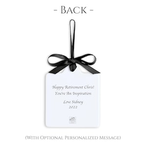 May include: A white, rectangular ornament with a black ribbon and bow. The ornament has the text "Happy Retirement Chris! You're An Inspiration Love Sidney 2022". The words "- BACK -" are at the top.