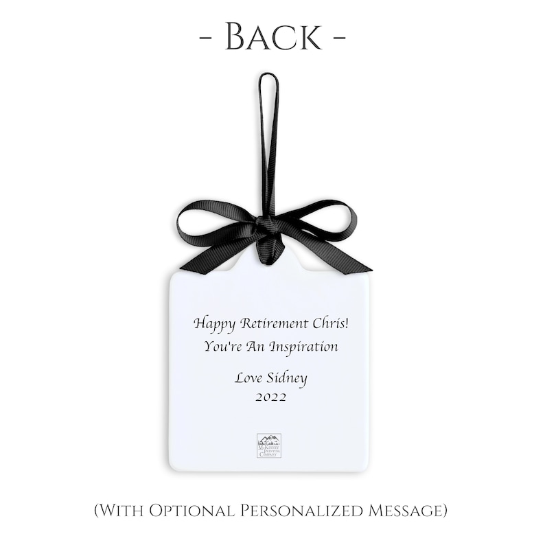 May include: A white, rectangular ornament with a black ribbon and bow. The ornament has the text "Happy Retirement Chris! You're An Inspiration Love Sidney 2022". The words "- BACK -" are at the top.