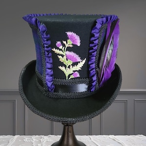 May include: A black top hat featuring a purple ruffled trim along the sides and a central embroidered thistle design in shades of pink and green. The hat includes a black satin band, decorative black braided trim, and a purple feather accent.
