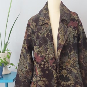 Op de afbeelding: Een lange, bruine, groene en gouden gepatroneerde jas met een shawlkraag en een enkele knoopsluiting. De jas is op een mannequin en heeft een licht uitlopende zoom.