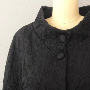 May include: A black, long-sleeved cape with a high collar and button closure. The cape is made of a textured fabric with a floral pattern.