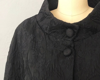 May include: A black, long-sleeved cape with a high collar and button closure. The cape is made of a textured fabric with a floral pattern.