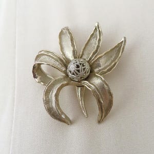 May include: A silver-toned flower brooch with a textured, openwork center and curved petals. The brooch is pinned to a cream-colored fabric, showcasing its elegant design. The flower's petals have a subtle, reflective sheen.