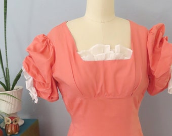 Könnte beinhalten: Ein Vintage-Kleid in Korallenrosa mit einer weißen Spitzenborte am Ausschnitt und kurzen, bauschigen Ärmeln. Das Kleid hat ein tailliertes Oberteil und einen weiten Rock.