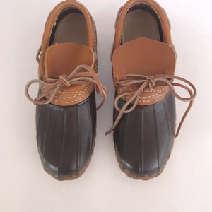 Könnte beinhalten: Ein Paar braune Leder- und Gummistiefel mit braunen Schnürsenkeln. Die Stiefel haben ein braunes Leder-Oberteil und ein schwarzes Gummi-Unterteil. Die Schnürsenkel sind oben an den Stiefeln zu einer Schleife gebunden.
