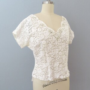 May include: White lace top with a V-neckline and short sleeves. The top is made of delicate lace with a floral pattern.