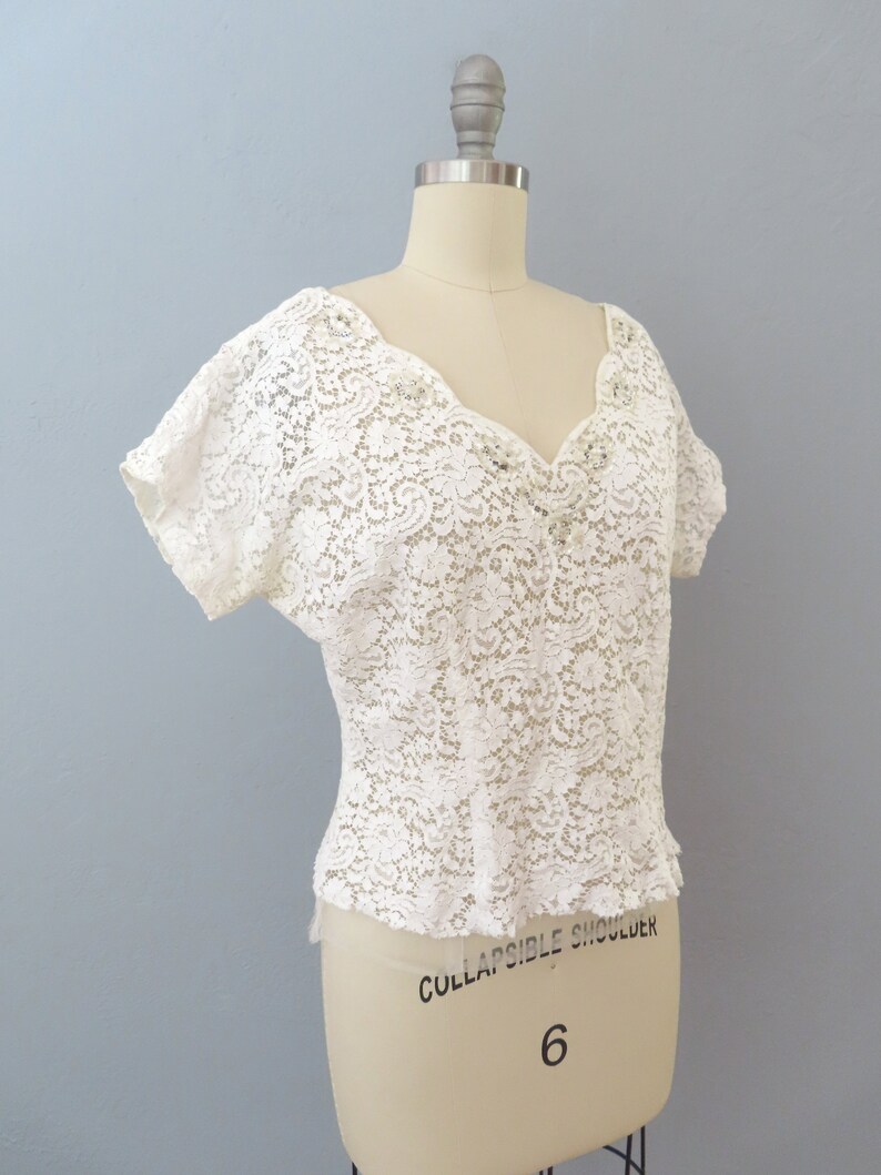 May include: White lace top with a V-neckline and short sleeves. The top is made of delicate lace with a floral pattern.