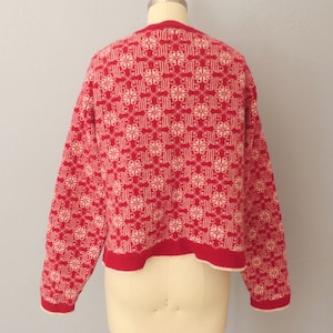 May include: Red knitted sweater with a white geometric pattern. The sweater has a crew neck and long sleeves.