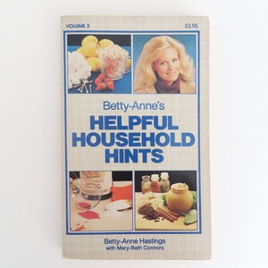 Peut inclure: Couverture d'un livre vintage intitul&eacute; "Betty-Anne's Helpful Household Hints, Volume 2". La couverture pr&eacute;sente des images d'aliments, d'une femme et d'objets m&eacute;nagers. Le titre est en bleu, et le nom de l'auteur est &eacute;galement visible.