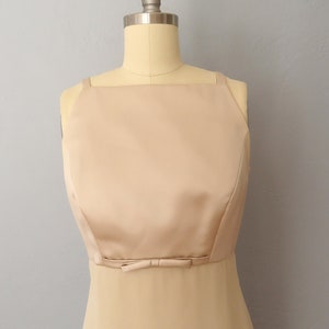 May include: A sleeveless, floor-length dress in a pale beige colour. The dress has a square neckline and a bow at the waist.