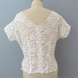 May include: White lace top with a V-neck and short sleeves. The top is made of delicate lace with a floral pattern.