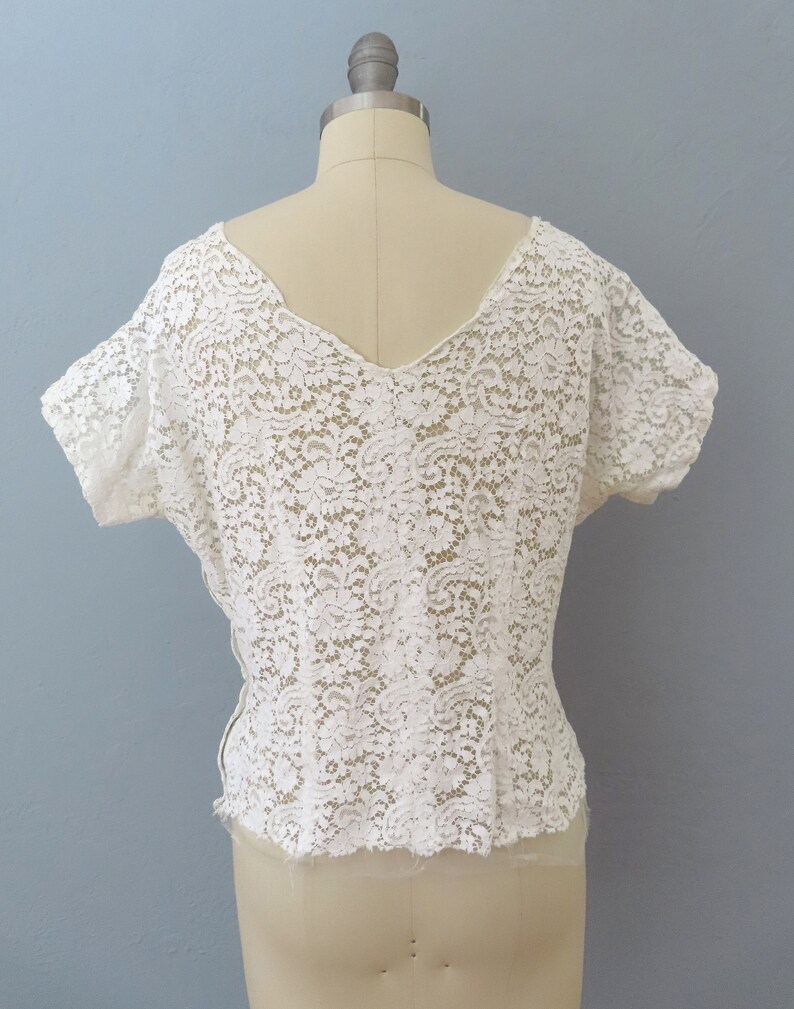 May include: White lace top with a V-neck and short sleeves. The top is made of delicate lace with a floral pattern.