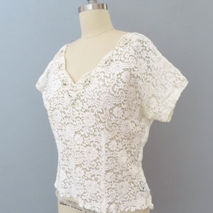 May include: White lace top with a V-neckline and short sleeves. The top has a delicate floral pattern and is fitted.
