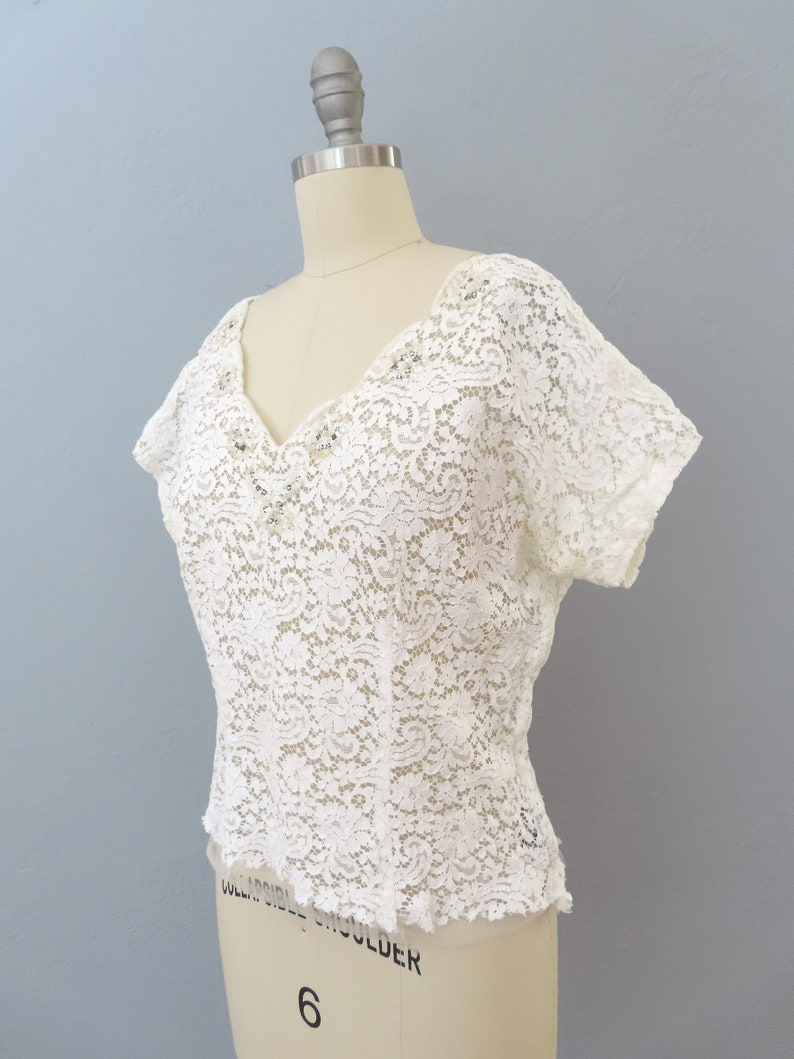 May include: White lace top with a V-neckline and short sleeves. The top has a delicate floral pattern and is fitted.