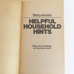 Peut inclure: Livre ouvert pr&eacute;sentant le titre "Betty-Anne's Helpful Household Hints" en gras noir. Les noms des auteurs, Betty-Anne Hastings et Mary-Beth Connors, sont imprim&eacute;s en dessous. Les pages sont jaunies.