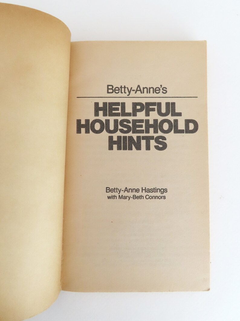 Peut inclure: Livre ouvert pr&eacute;sentant le titre "Betty-Anne's Helpful Household Hints" en gras noir. Les noms des auteurs, Betty-Anne Hastings et Mary-Beth Connors, sont imprim&eacute;s en dessous. Les pages sont jaunies.