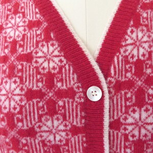 May include: Red cardigan with a white snowflake pattern and white trim. The cardigan has two white buttons.