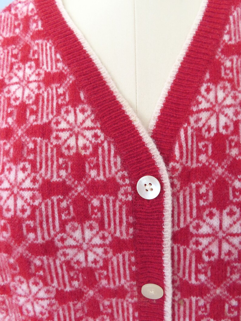 May include: Red cardigan with a white snowflake pattern and white trim. The cardigan has two white buttons.
