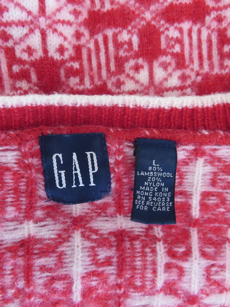 May include: A red and white sweater with a geometric pattern. The sweater is labeled "GAP" and has a size tag that reads "L 80% Lambswool 20% Nylon Made in Hong Kong RN 54023 See Reverse for Care."