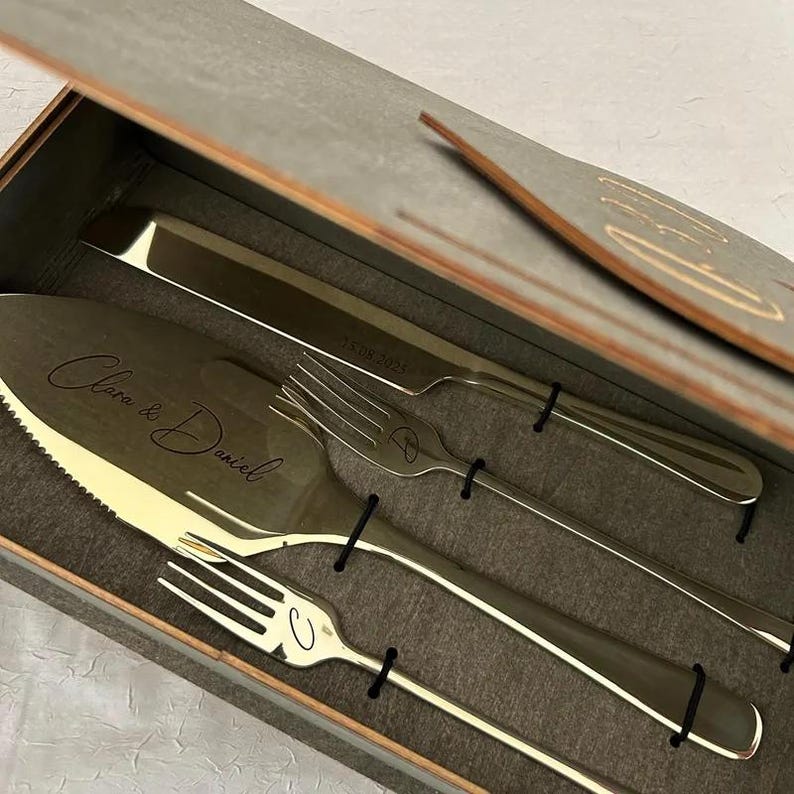 Personalized 4-piece champagne gold wedding cake cutting set presented in a premium wooden gift box. The serrated cake server is custom engraved with the names "Clara & Daniel" in elegant script. The set includes a matching gold knife with the wedding date "15.08.2025" and two dessert forks featuring script initials "C" and "D". Each piece is secured within a grey foam insert, showcasing a luxury unboxing experience for a bride and groom or as a wedding anniversary gift.