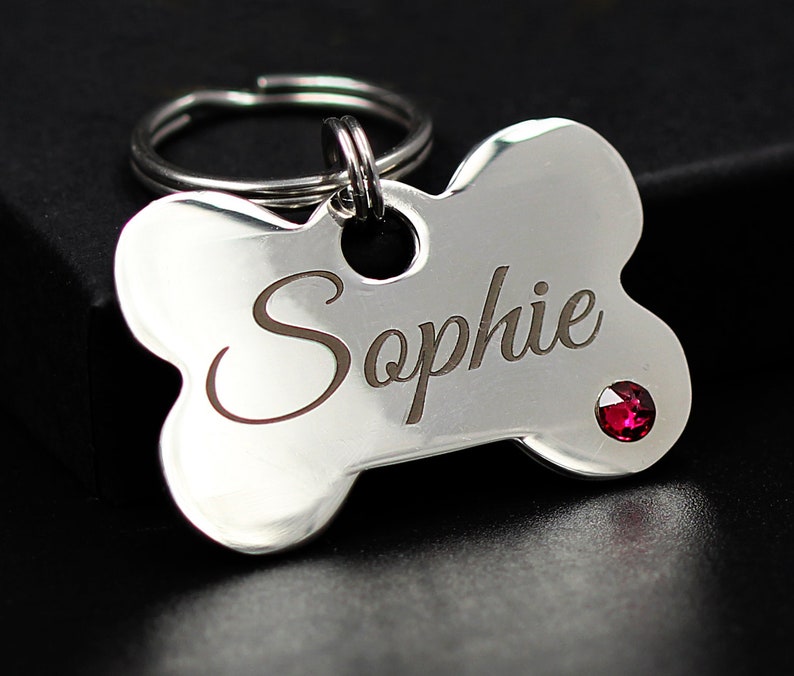 May include: Silver bone-shaped dog tag with the name "Sophie" engraved on it. The tag has a small red rhinestone.