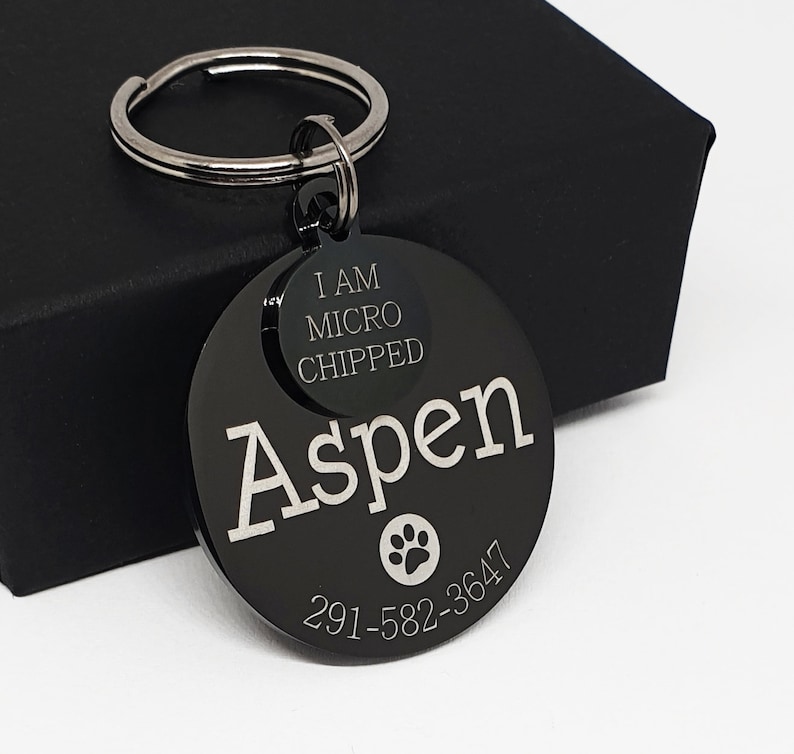 May include: Black metal dog tag with the name Aspen engraved on it. The tag also includes the text "I am microchipped" and a phone number.