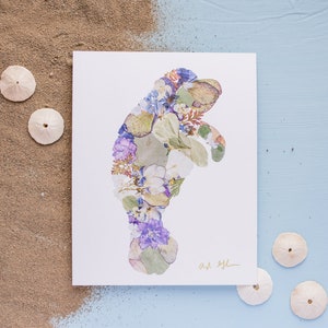 May include: A white card with a manatee silhouette made of pressed flowers and leaves. The flowers are in shades of purple, blue, white, and green. The card is signed "Art by L." and is surrounded by sand dollars on a blue background.