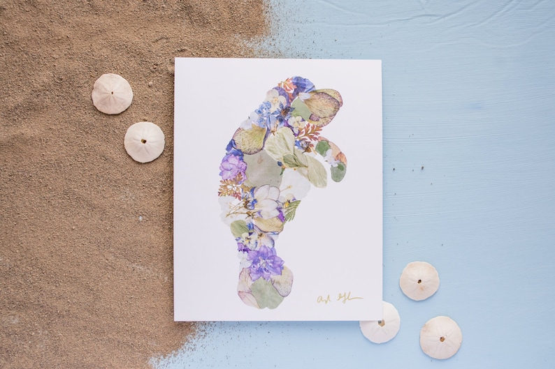 May include: A white card with a manatee silhouette made of pressed flowers and leaves. The flowers are in shades of purple, blue, white, and green. The card is signed "Art by L." and is surrounded by sand dollars on a blue background.
