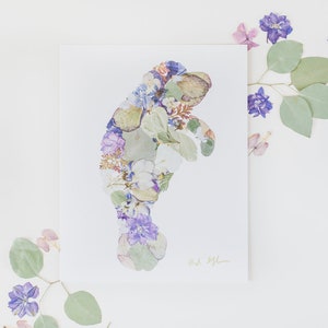May include: A print of a manatee made with pressed flowers and leaves. The manatee is outlined in white and filled with purple, white, and green flowers. The print is signed "Art by Jill".