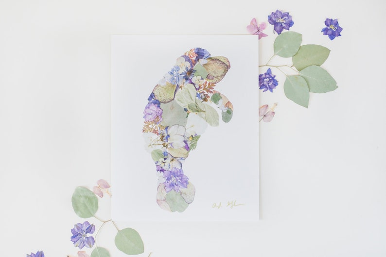May include: A print of a manatee made with pressed flowers and leaves. The manatee is outlined in white and filled with purple, white, and green flowers. The print is signed "Art by Jill".